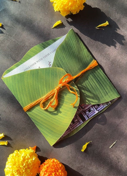 A paper invitation card designed to look like a banana leaf, tied with an orange thread, displayed on a surface with floral decorations around it.